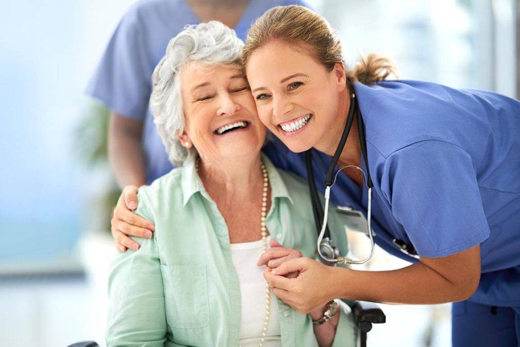 Photo of a smiling older women being hugged by a happy healthcare provider.